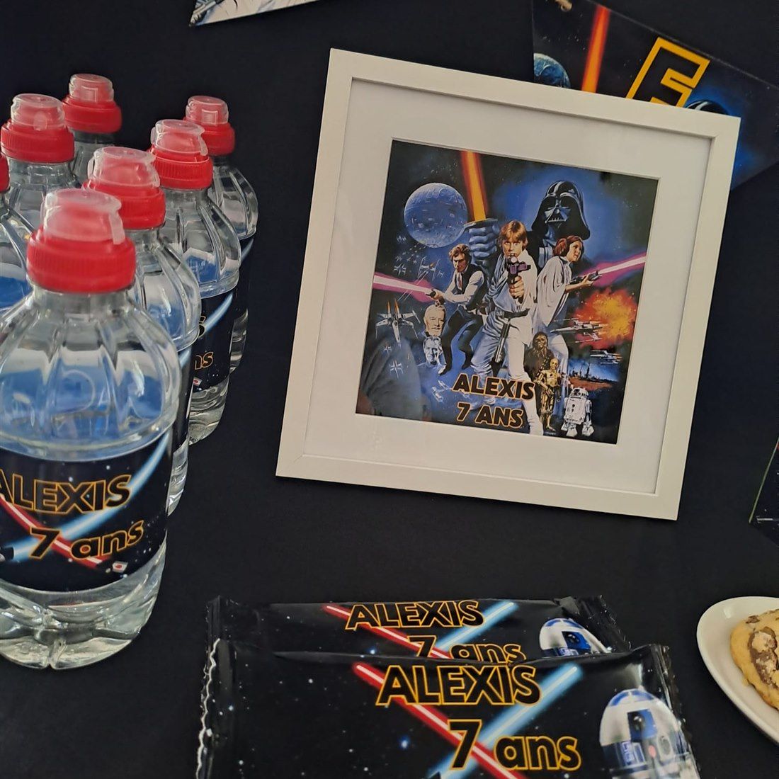 Table d'anniversaire sur un thème galactique : bouteilles d'eau, sachets de gâteaux, guirlande de fanions et cadre souvenir, tous personnalisés.