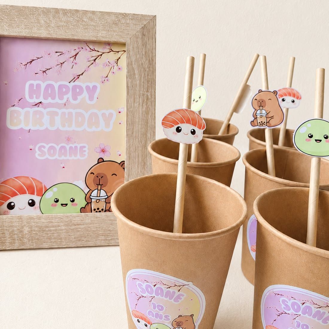 Gobelets en kraft avec pailles décorées de personnages kawaï (sushi, capybara, mochi) et cadre « Happy Birthday » aux tons pastel.