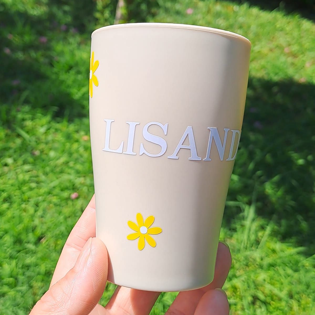 Gobelet beige au prénom Lisandro en lettres blanches, décoré de marguerites jaunes, tenu au soleil au-dessus d'un set en raphia.