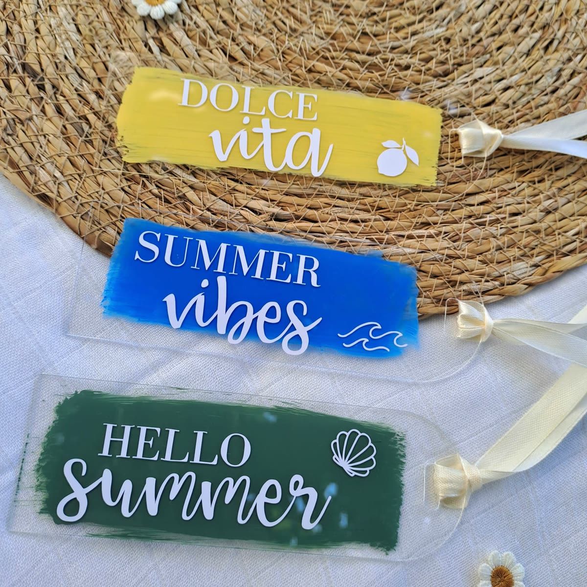 Trois marque-pages en plexi transparent peints à la main et noués d'un ruban ivoire : un jaune « Dolce vita » avec un citron, un bleu « Summer vibes » avec des vagues, un vert foncé « Hello summer » avec un coquillage.