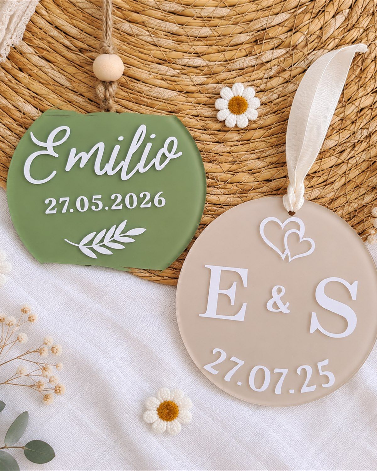 Deux ornements en plexi posés sur du raphia et un tissu blanc : un ornement vert sauge au prénom Emilio et à la date 27.05.2026 orné d'une branche, et un ornement rond beige aux initiales E & S surmontées de deux cœurs, daté 27.07.25.