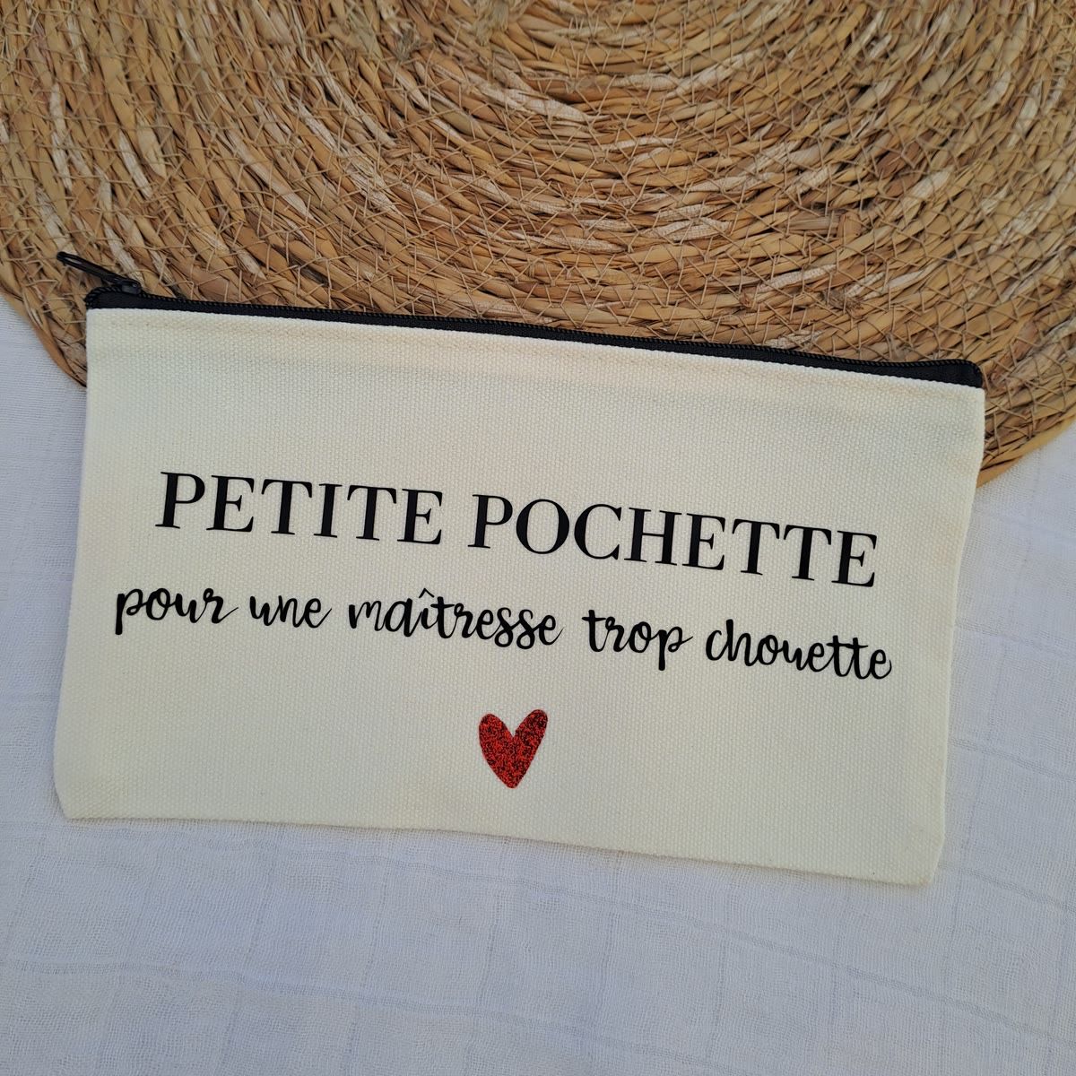 Pochette en toile écrue à fermeture éclair noire, floquée « Petite pochette pour une maîtresse trop chouette » et d'un petit cœur rouge, posée sur un set en raphia.