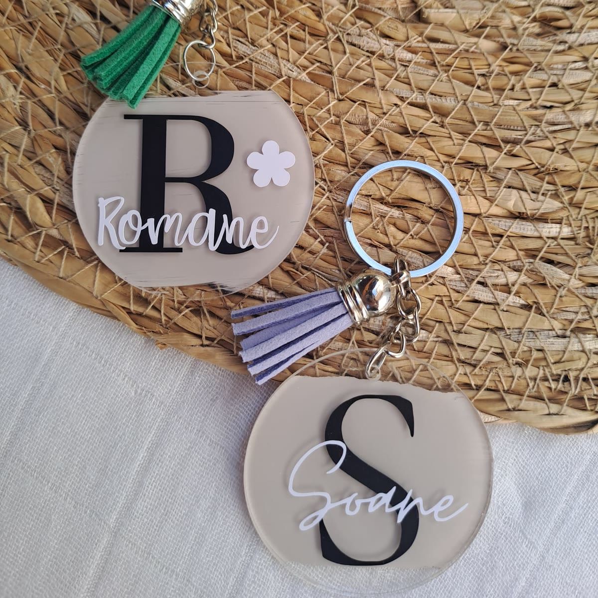 Deux porte-clés ronds en acrylique sur un set en raphia : l'un à l'initiale R et au prénom Romane avec un pompon vert, l'autre à l'initiale S et au prénom Soane avec un pompon lilas.