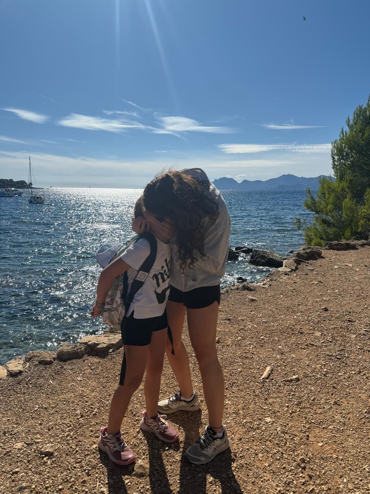 Floriane et sa fille Soane enlacées, de dos, sur un chemin de bord de mer, face à une crique ensoleillée.